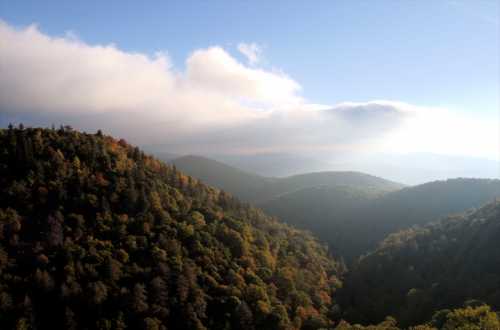 A scenic view of rolling green mountains under a blue sky with soft clouds, showcasing autumn foliage.