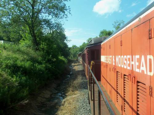 A train car labeled "East & Moosehead" travels along a green, tree-lined track beside a small stream.