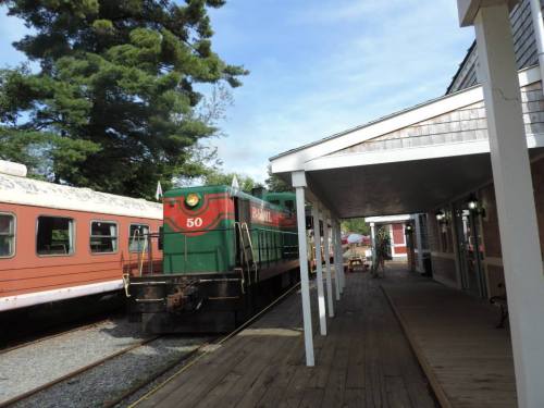A green and red train engine parked beside a wooden platform, with trees and a vintage train car in the background.