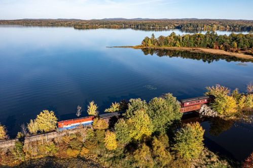 A train travels along a scenic lakeside, surrounded by colorful autumn foliage and calm waters.