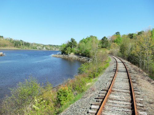 A scenic view of a winding railway track alongside a calm river, surrounded by lush greenery and clear blue skies.