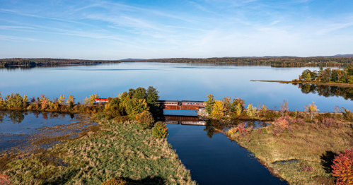 Aerial view of a tranquil lake surrounded by autumn foliage and a train crossing a bridge over calm waters.