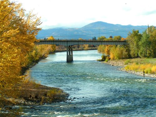 A river flows under a bridge, surrounded by autumn trees and distant mountains under a clear sky.