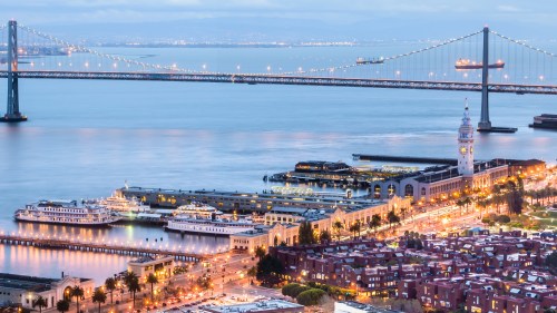 A scenic view of a waterfront city with a bridge, illuminated buildings, and boats on the water at dusk.