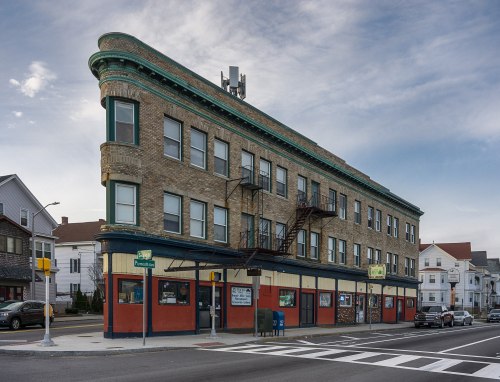 Historic brick building with a curved facade, featuring large windows and a ground-level storefront, set on a street corner.