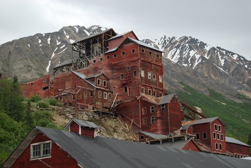 Abandoned red mining buildings on a hillside, surrounded by green vegetation and snow-capped mountains in the background.