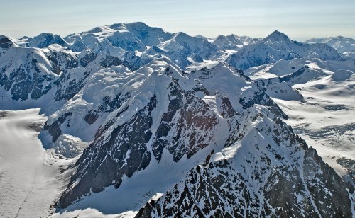 Aerial view of snow-covered mountains with rugged peaks and glaciers under a clear blue sky.