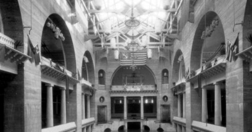 Historic interior of a grand hall with arches, flags, and a large skylight, showcasing architectural details.