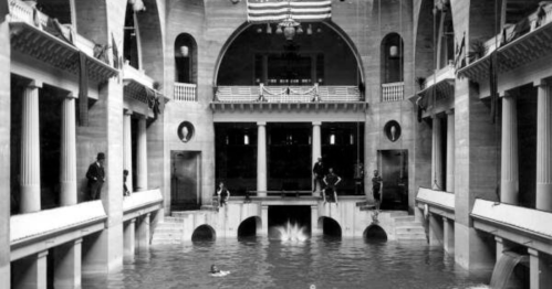 Historic indoor swimming pool with columns, flags, and people swimming in a flooded space. Black and white photo.