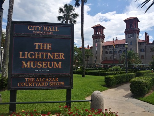 Sign for the Lightner Museum in front of City Hall, featuring palm trees and a historic building in the background.