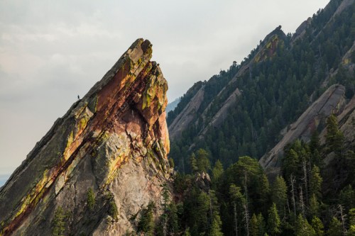 A jagged rock formation rises above a forested landscape, with mountains in the background under a cloudy sky.