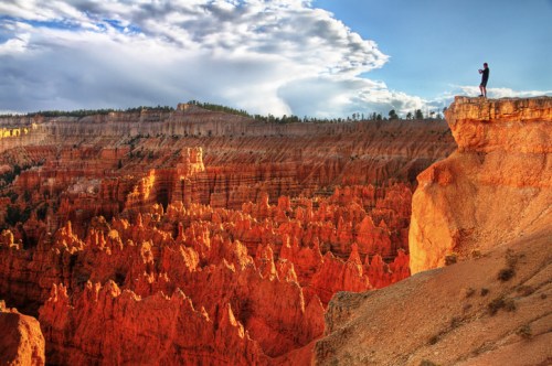 A person stands on a cliff overlooking the vibrant orange rock formations of Bryce Canyon under a cloudy sky.