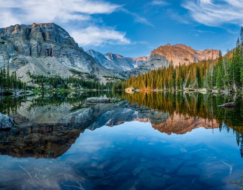 A serene mountain lake reflecting towering peaks and lush pine trees under a clear blue sky.