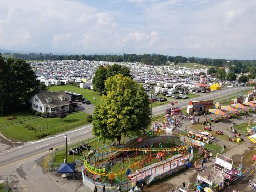 Aerial view of a fairground with rides, food stalls, and a large parking area filled with vehicles in the background.