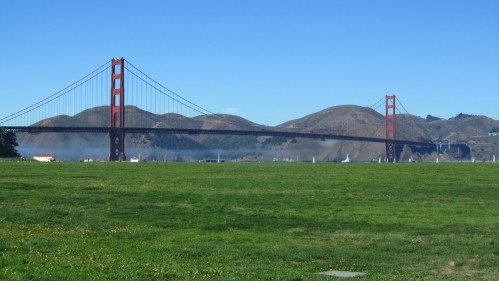 Golden Gate Bridge spans across a bay, with green grass in the foreground and hills in the background under a clear blue sky.