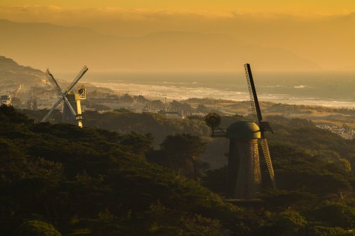 Two windmills stand amidst trees overlooking a coastline at sunset, with mountains in the background and soft golden light.