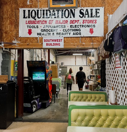 A sign for a liquidation sale hangs above a corridor with green booths and shoppers walking towards the back.