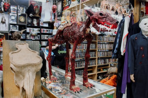 A grotesque, bloodied creature stands on display in a cluttered horror-themed shop filled with masks and DVDs.