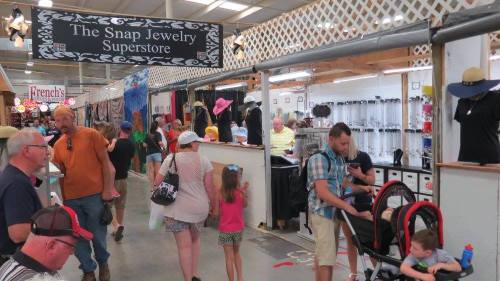 A busy market scene with shoppers browsing jewelry and clothing stalls, including families and children.