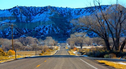A winding road leads through a snowy landscape with mountains and bare trees under a clear blue sky.