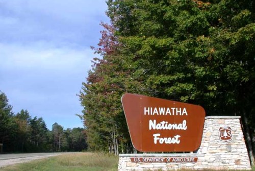 Sign for Hiawatha National Forest, surrounded by trees with autumn foliage, along a road under a partly cloudy sky.