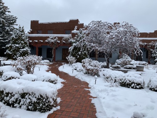 A snowy landscape featuring a brick pathway leading to a building surrounded by snow-covered shrubs and trees.