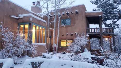 A snow-covered adobe house with large windows, surrounded by trees and a winter landscape.