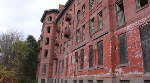Abandoned brick building with peeling paint, broken windows, and fire escapes, surrounded by overgrown vegetation.