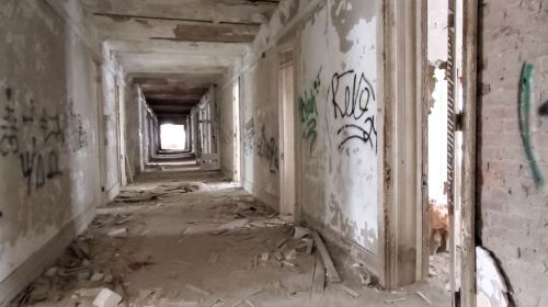 Abandoned hallway with peeling walls, graffiti, and debris scattered on the floor, leading to a bright exit at the end.