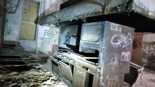 Abandoned kitchen with old, rusty appliances and graffiti on the walls, illuminated by dim light.