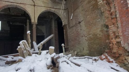 Snow-covered debris and rubble inside a dilapidated building with brick walls and arched doorways.