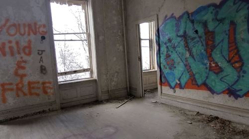 An abandoned room with peeling walls, two windows, and colorful graffiti on one wall. Dust and debris on the floor.