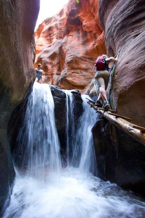 Two hikers navigate a rocky canyon with a waterfall, using a wooden bridge to cross the rushing water.