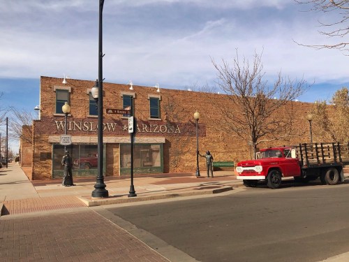 A red truck parked near a brick building with "Winslow Arizona" signage and a statue on the sidewalk.