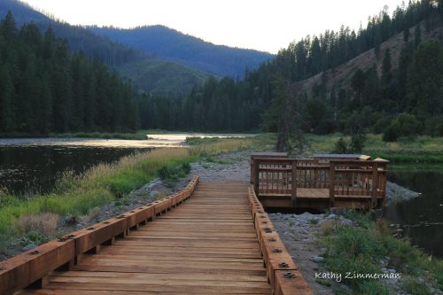 A wooden walkway leads to a scenic river surrounded by lush trees and mountains at dusk.