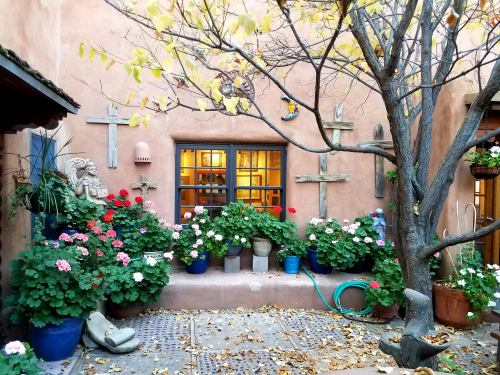 A charming courtyard with colorful flowers, a tree, and decorative crosses against a warm, adobe wall.