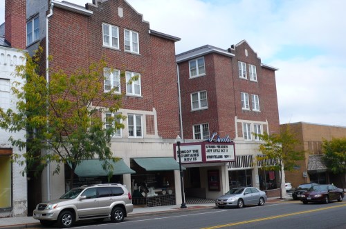 A brick building with a theater marquee and shops below, set on a street with parked cars and trees.
