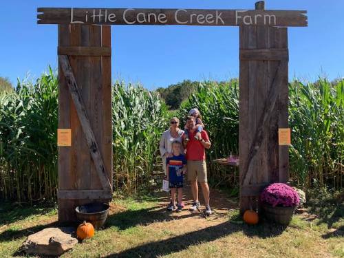 A family stands at the entrance of Little Cane Creek Farm, surrounded by cornfields and autumn decorations.