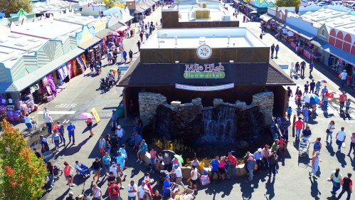 Aerial view of a bustling flea market with colorful stalls and a waterfall feature, filled with people shopping.