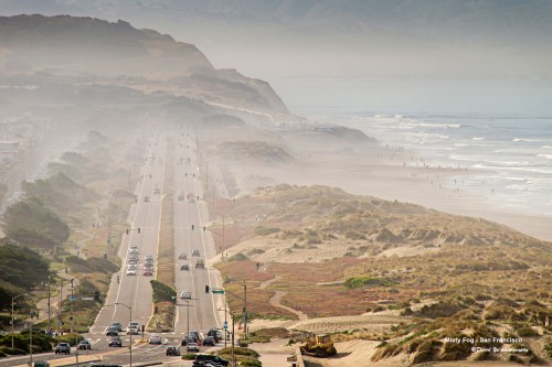 A coastal road winds through foggy hills, leading to a beach with gentle waves and distant figures enjoying the shore.
