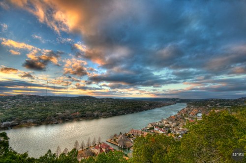 A scenic view of a river winding through lush greenery, with colorful clouds at sunset and houses along the shore.