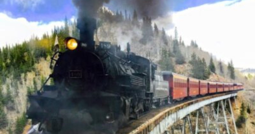 A vintage steam train crosses a wooden trestle bridge surrounded by lush green trees and a cloudy sky.