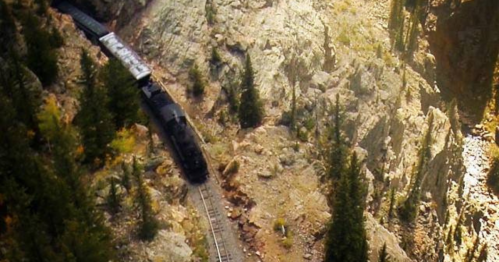 A train travels along a winding track through rocky terrain and trees, surrounded by a mountainous landscape.