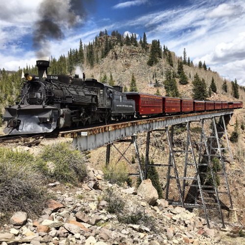 A vintage steam train crosses a tall bridge surrounded by trees and mountains under a blue sky with clouds.