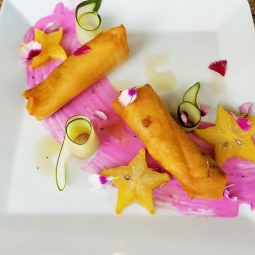 Two spring rolls on a colorful plate with pink sauce, garnished with star fruit, edible flowers, and green onion curls.