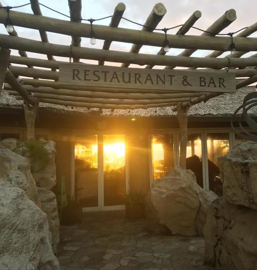 A rustic restaurant and bar entrance with a wooden sign, surrounded by rocks and plants, glowing in the sunset.