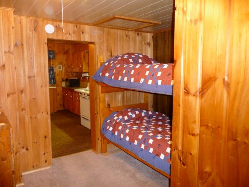 A cozy room with wooden walls featuring a set of bunk beds and a view into a kitchen area.