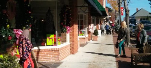 A festive street scene with holiday decorations, shoppers, and storefronts adorned with wreaths and gifts.