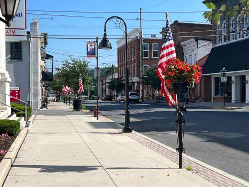 A quiet street lined with American flags and flower baskets, featuring shops and lampposts under a clear blue sky.
