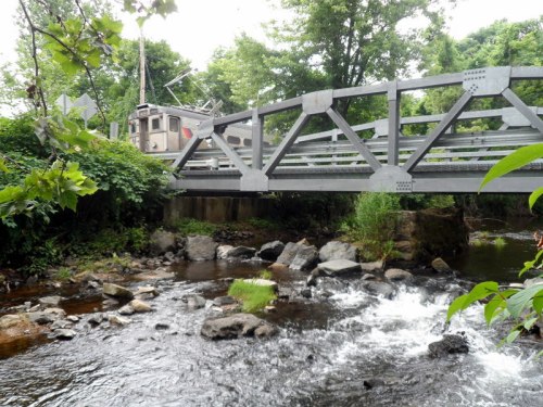 A metal bridge spans a small stream, surrounded by lush greenery and rocky banks.